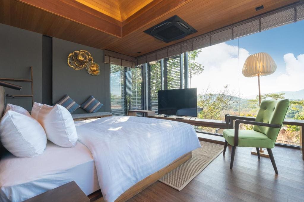 Modern bedroom with large windows, a neatly made bed, green armchair, floor lamp, TV, and scenic views of mountains and trees outside. Room features wood accents, soft lighting, and contemporary decor.