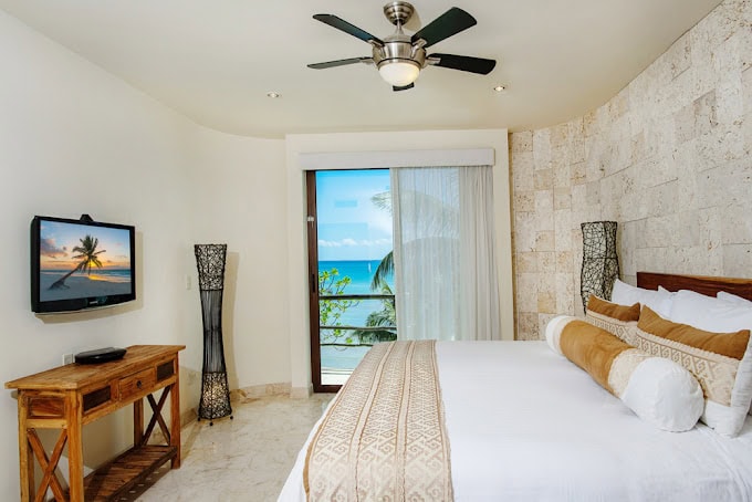 A bright hotel room features a large bed with tan pillows, a ceiling fan, a flat-screen TV on a wooden desk, two decorative lamps, and a balcony with an ocean view through sliding glass doors.