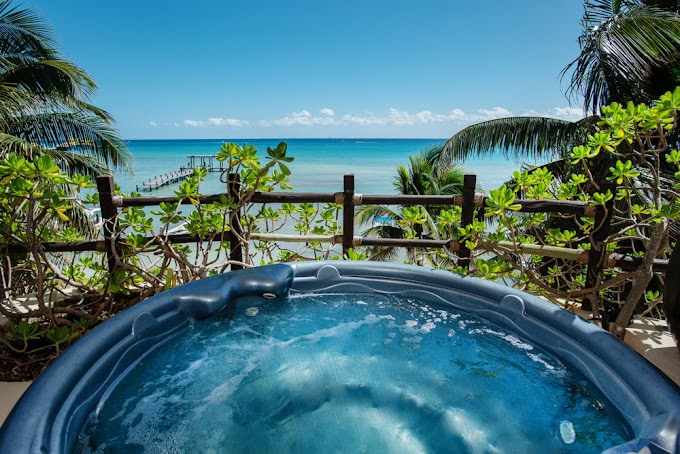 A bubbling outdoor hot tub overlooks a tropical beach with turquoise water, palm trees, green foliage, and a wooden pier extending into the ocean under a clear blue sky.