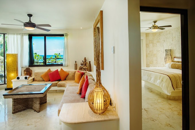 Bright, modern suite with a living area featuring a beige sectional sofa, colorful pillows, wicker table, and decor. Large windows show ocean views. A bedroom with neutral tones and a ceiling fan is visible through an open doorway.