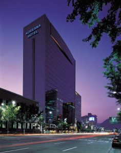 A tall, modern building labeled Koreana Hotel stands along a city street at dusk, with illuminated windows, light trails from passing cars, and trees lining the street under a purple sky.