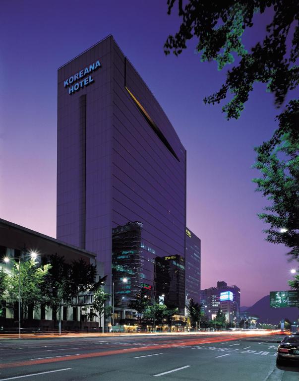 A tall, modern building labeled Koreana Hotel stands along a city street at dusk, with illuminated windows, light trails from passing cars, and trees lining the street under a purple sky.