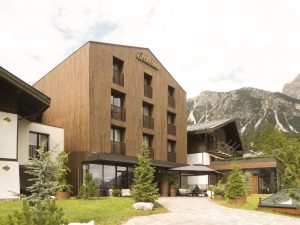 A modern, three-story wooden hotel building labeled “Faloria” is surrounded by greenery and mountains, with large windows and small balconies on each floor under a partly cloudy sky.