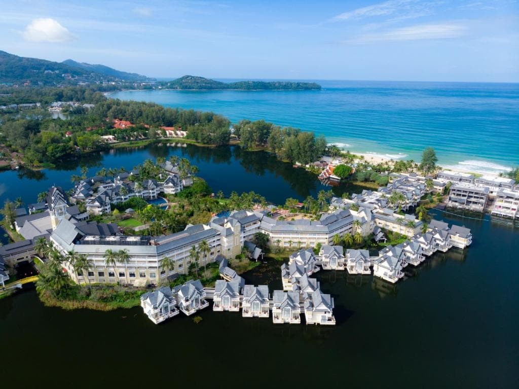 Aerial view of a coastal resort with white buildings surrounded by a lake and lush greenery, near a sandy beach and turquoise ocean. Hills and more greenery stretch into the background under a partly cloudy sky.