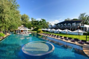 A large outdoor swimming pool with a circular spa, surrounded by lounge chairs and white umbrellas, sits next to a modern hotel building and lush greenery under a clear blue sky.