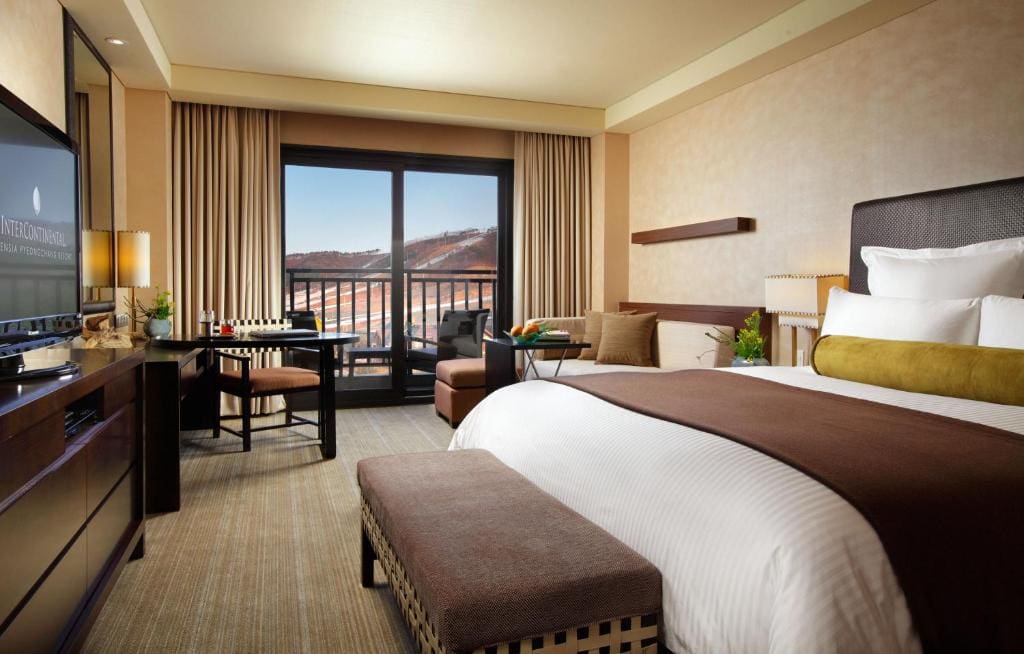 A modern hotel room with a large bed, TV, desk, and seating area. There are floor-to-ceiling windows with curtains, leading to a balcony with a view of hills or mountains in the distance.