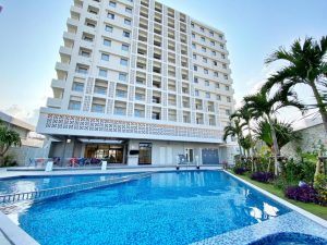 A modern, multi-story white apartment building with balconies overlooks a swimming pool surrounded by palm trees and greenery on a sunny day. Outdoor seating is visible near the pool area.