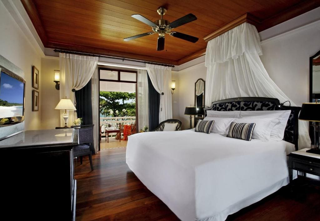 A stylish hotel room with a large bed, white linens, canopy, ceiling fan, wooden floors, a desk, lamps, and a balcony with outdoor seating overlooking greenery.