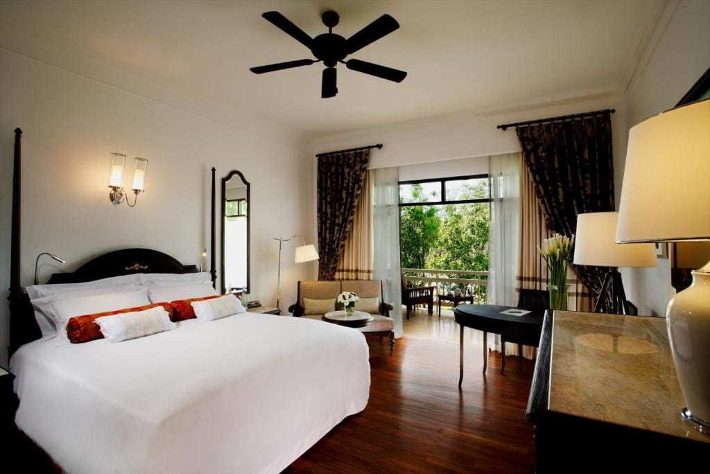 A modern hotel room with a large bed, white bedding, and decorative pillows. There is a ceiling fan, bedside lamps, a desk, a sofa, and large windows with curtains overlooking a balcony and greenery outside.