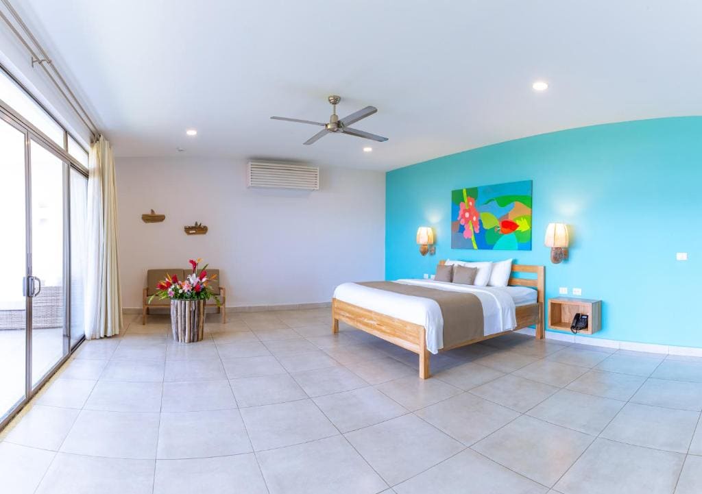 Spacious, modern bedroom with a king bed, turquoise accent wall, colorful artwork, bedside lamps, large windows with curtains, a chair, a ceiling fan, and a vase of flowers on a side table.