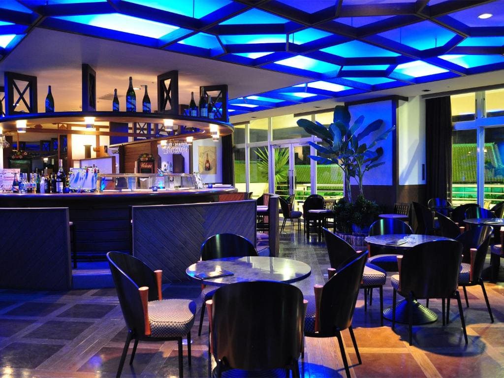 A modern bar with round tables, black chairs, and a curved counter. Blue ceiling lights create a vibrant atmosphere. Wine bottles decorate the counter, and large windows reveal greenery outside. The bar area is empty.