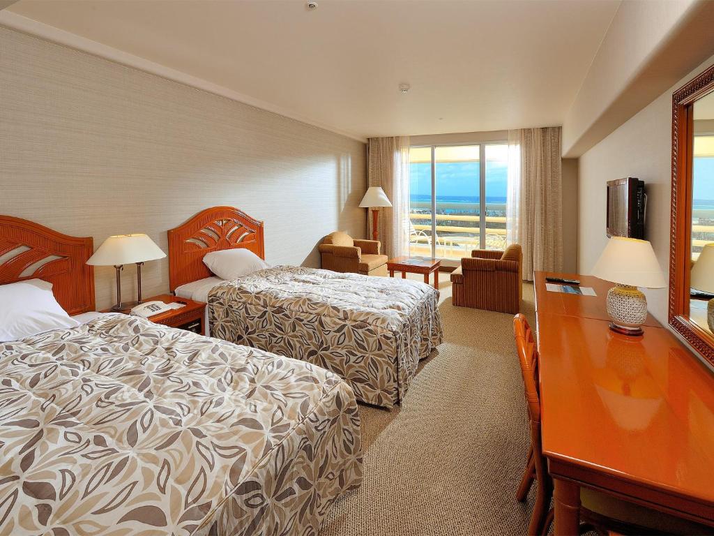 Hotel room with two twin beds, patterned bedspreads, wooden headboards, a desk with a mirror, TV, two armchairs by a window, lamps, and a view of blue sky and landscape outside.