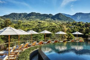 Lounge chairs and white umbrellas line a curved infinity pool overlooking lush green hills and distant mountains under a clear blue sky.