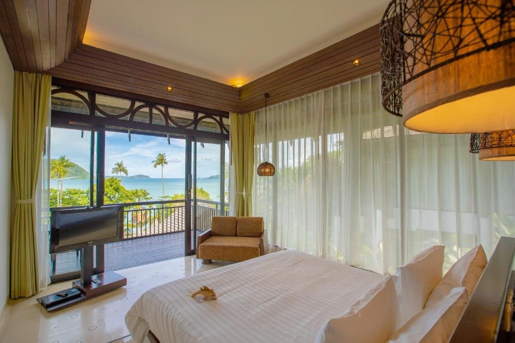 A bright, modern bedroom with a large bed, glass walls with sheer curtains, a lounge chair, and a TV. The room opens to a balcony with views of palm trees, greenery, and the ocean under a clear sky.