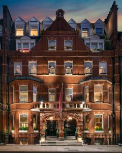 A grand, red-brick Victorian building at dusk, featuring ornate architectural details, large windows with warm lights inside, and a maroon flag above two arched entrances flanked by columns and greenery.