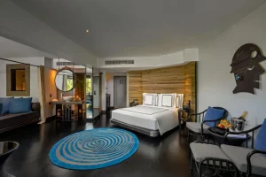 Modern hotel room with a large bed, blue circular rug, seating area with two chairs and a table, wooden accent wall, open bathroom area, and minimalistic decor with natural light coming in.