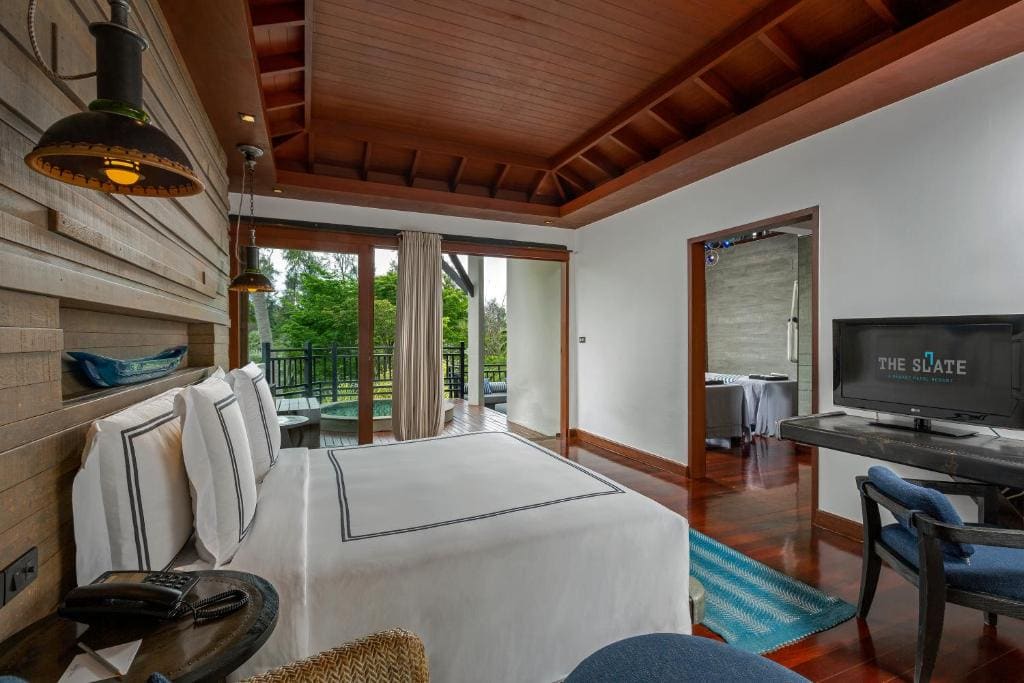 A modern hotel room with a large bed, wooden ceiling, desk with TV, glass doors opening to a balcony with greenery outside, and a stylish, minimal decor.