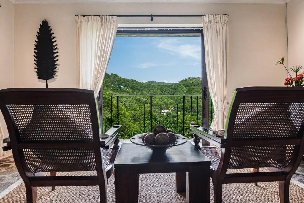 Two wooden chairs face a small table with a decorative bowl, set by a window with white curtains. The window opens to a balcony overlooking lush green hills under a blue sky.