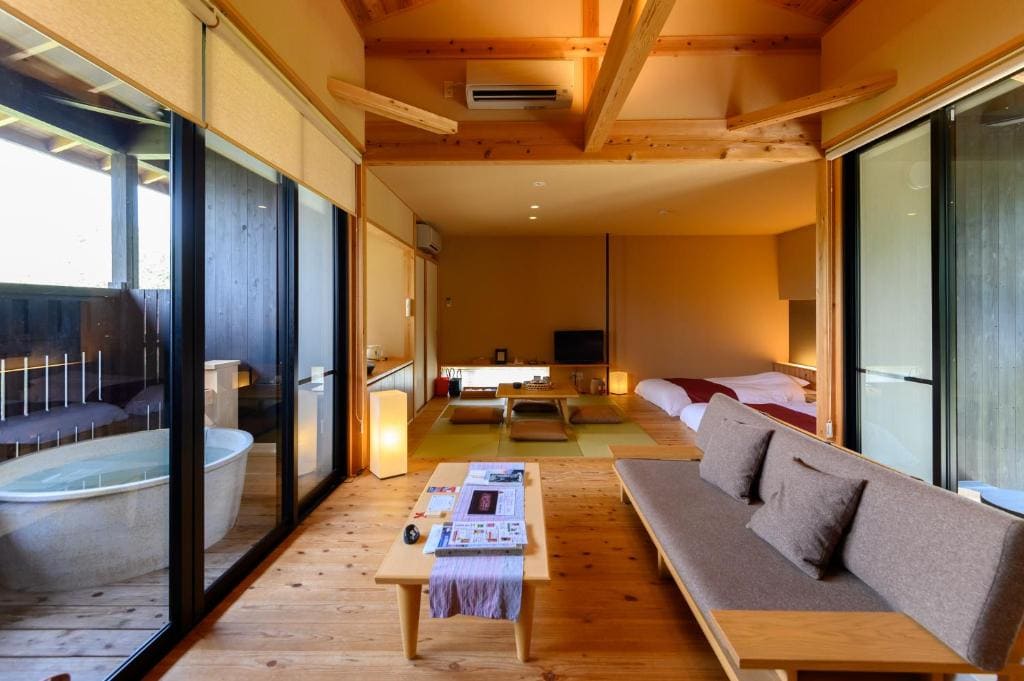 A modern Japanese-style room with wooden floors, a sofa, and a low table in the living area. Sliding glass doors open to a patio with a bathtub. Futon beds and a tatami mat dining area are in the background. Natural light fills the space.