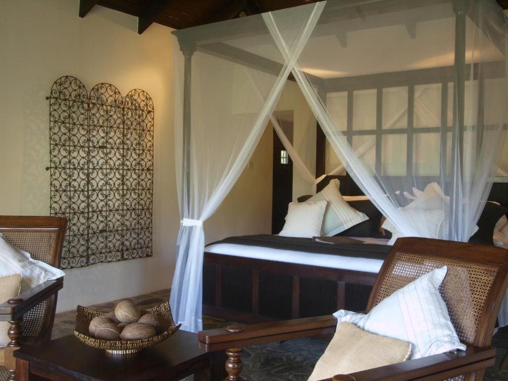 A cozy bedroom with a four-poster bed draped in white mosquito netting, wooden chairs with cushions, a bowl of decorative items on a table, and a decorative wrought iron wall piece. The room has warm, inviting lighting.