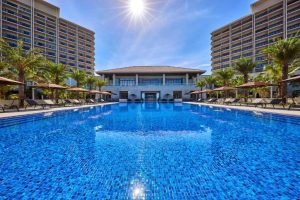 A sparkling blue outdoor pool is surrounded by lounge chairs, umbrellas, and palm trees, with a modern resort building and tall towers in the background under a sunny, clear sky.
