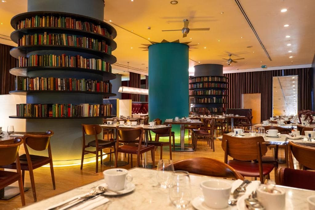 A cozy restaurant with round bookshelves built around columns, wooden chairs and tables set for dining, white cups and saucers, and warm lighting creating an inviting atmosphere.