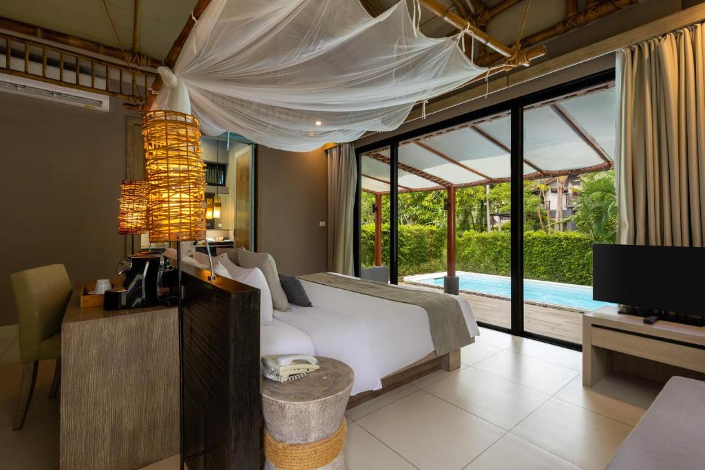 Modern hotel room with a large bed under a white mosquito net, a desk with a lamp, a TV, and glass doors opening to a private patio with a pool and greenery outside. The space feels bright and inviting.