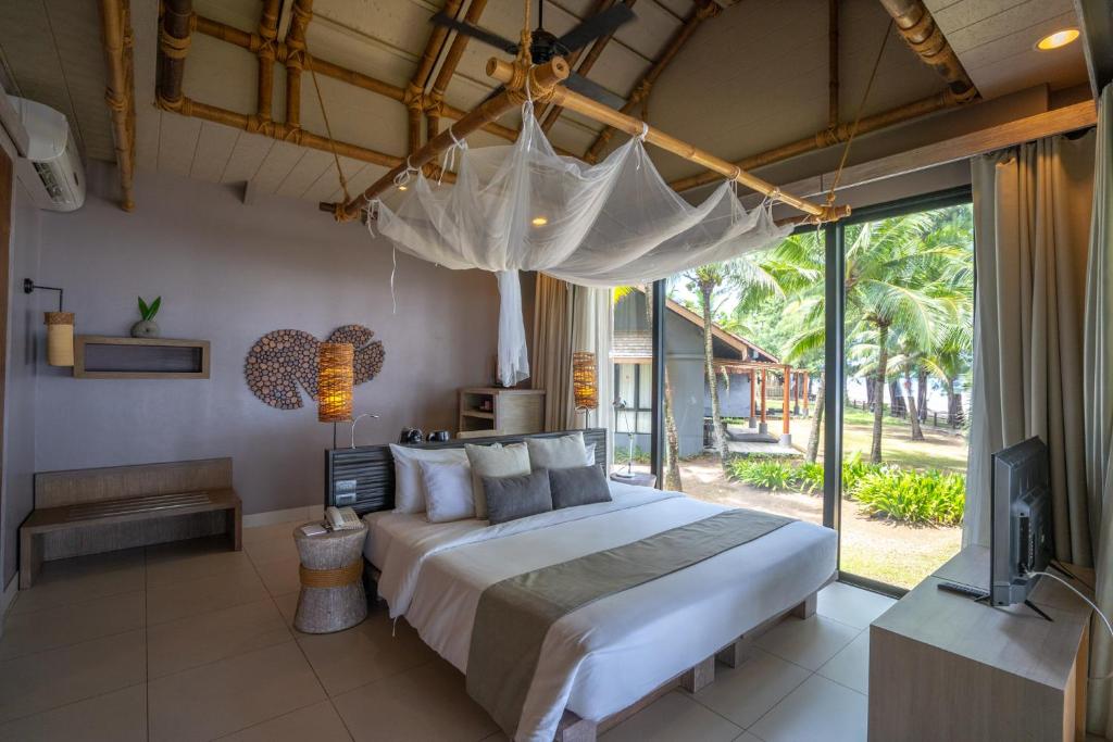 A modern tropical bedroom with a large bed under a white mosquito net, light wood furniture, and glass doors opening to a view of palm trees and a beachside hut. Warm, natural light fills the room.