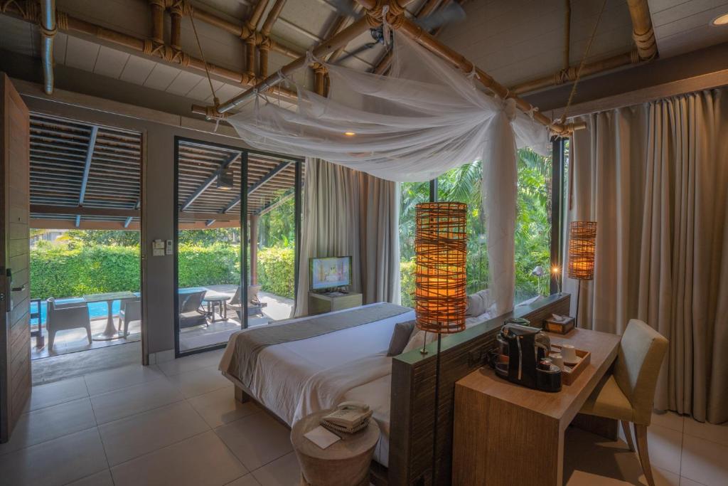 A cozy resort room with a canopy bed, desk, and coffee setup. Large windows reveal a private patio with lounge chairs, a small pool, and lush greenery outside. Warm, natural lighting fills the space.