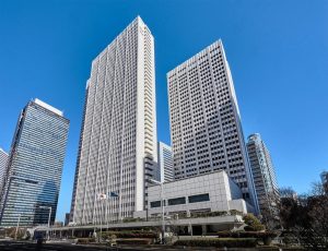 Tall modern office buildings with glass windows and geometric patterns stand against a clear blue sky. Flags are displayed in front of the main building, and trimmed trees and hedges line the entrance area.