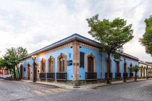 A corner building with bright blue walls, tall arched windows, and decorative iron railings sits on a cobblestone street lined with trees under a cloudy sky.