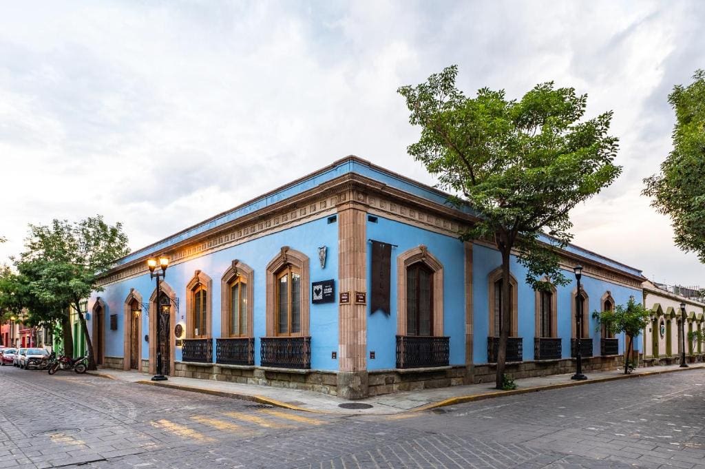 A corner building with bright blue walls, tall arched windows, and decorative iron railings sits on a cobblestone street lined with trees under a cloudy sky.