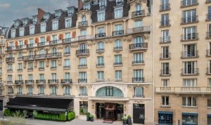 A large, elegant stone building with ornate balconies, arched windows, and a black awning over the entrance. The architecture features classic Parisian style, with mansard roofs and decorative detailing.