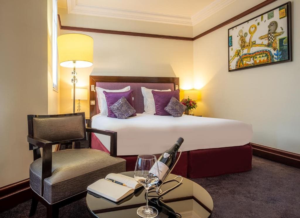 A neatly made hotel bed with purple cushions, a side lamp, and artwork above the headboard. In front, a round table holds a wine bottle, glass, notebook, and pen next to a cushioned chair.