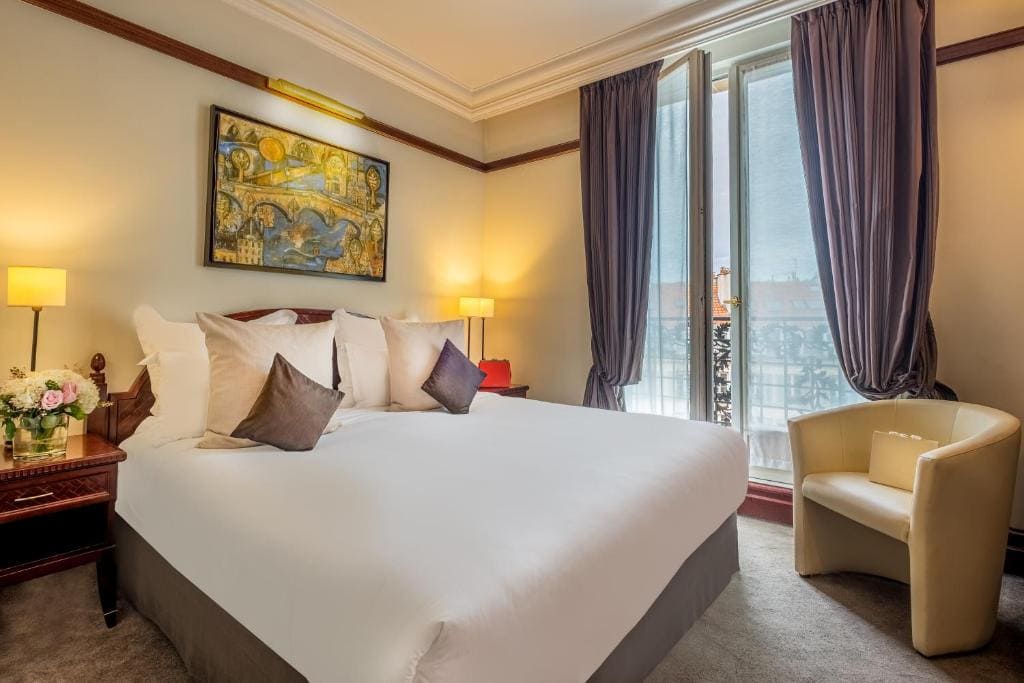 A cozy hotel room with a large bed, white linens, and several pillows, flanked by two bedside tables with lamps. A painting hangs above the bed, and tall windows with purple curtains let in daylight. A beige armchair sits near the window.