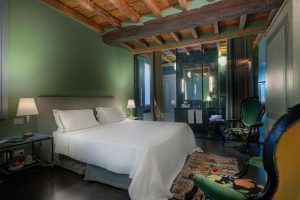 A cozy bedroom with green walls, a wooden beamed ceiling, a double bed with white linens, bedside lamps, and an ornate green armchair. An open bathroom area with mirrors is visible behind glass.