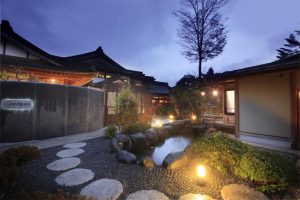 A serene Japanese garden at dusk with stone steps, a small pond, lantern lights, lush greenery, and traditional wooden buildings with tiled roofs in the background under a twilight sky.