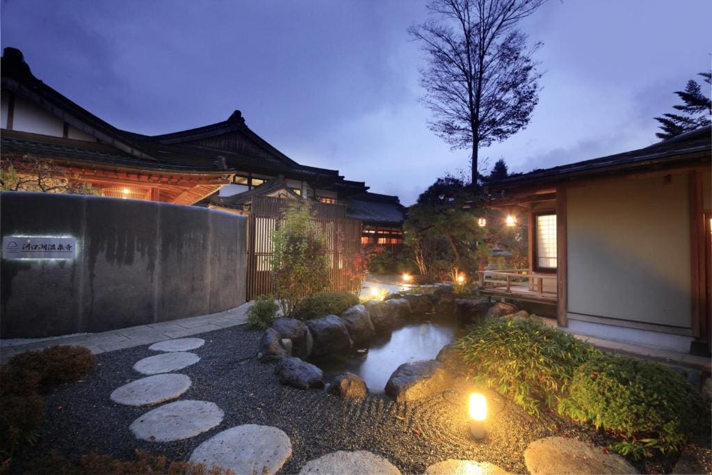 A serene Japanese garden at dusk with stone steps, a small pond, lantern lights, lush greenery, and traditional wooden buildings with tiled roofs in the background under a twilight sky.