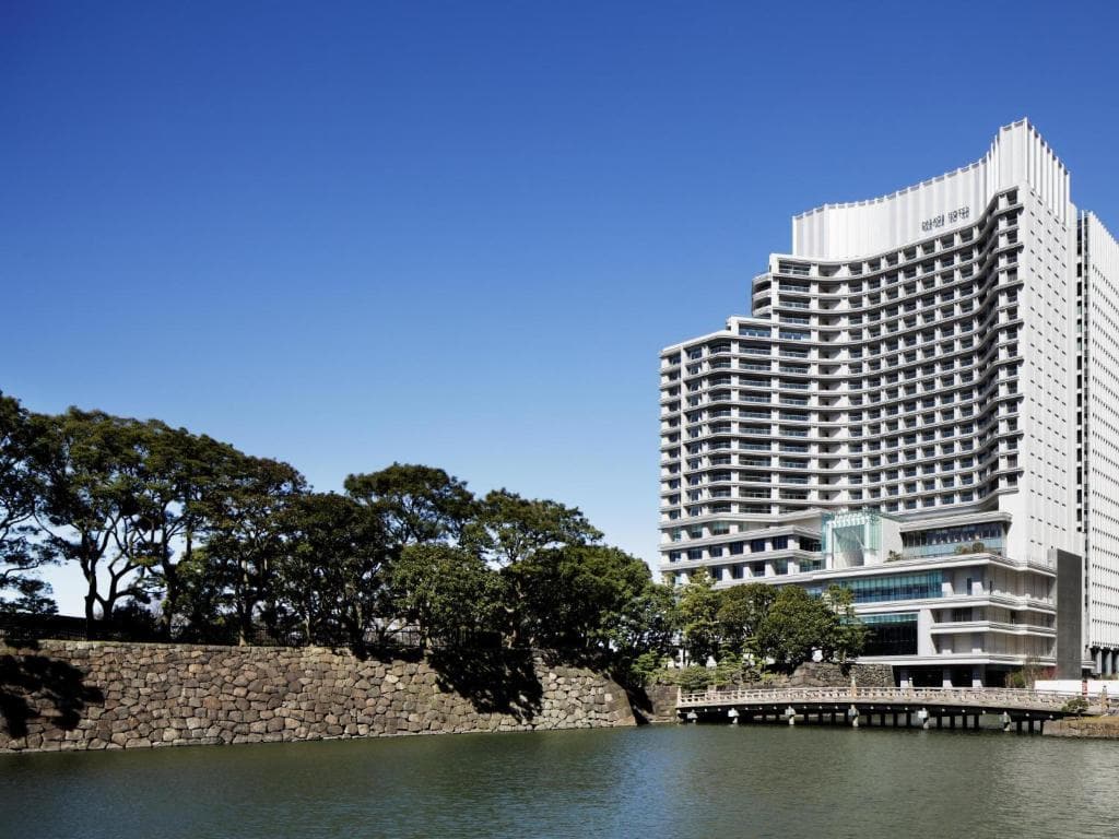 A tall, modern white hotel building stands beside a calm lake, with a wooden bridge and stone wall in the foreground and trees along the water under a clear blue sky.
