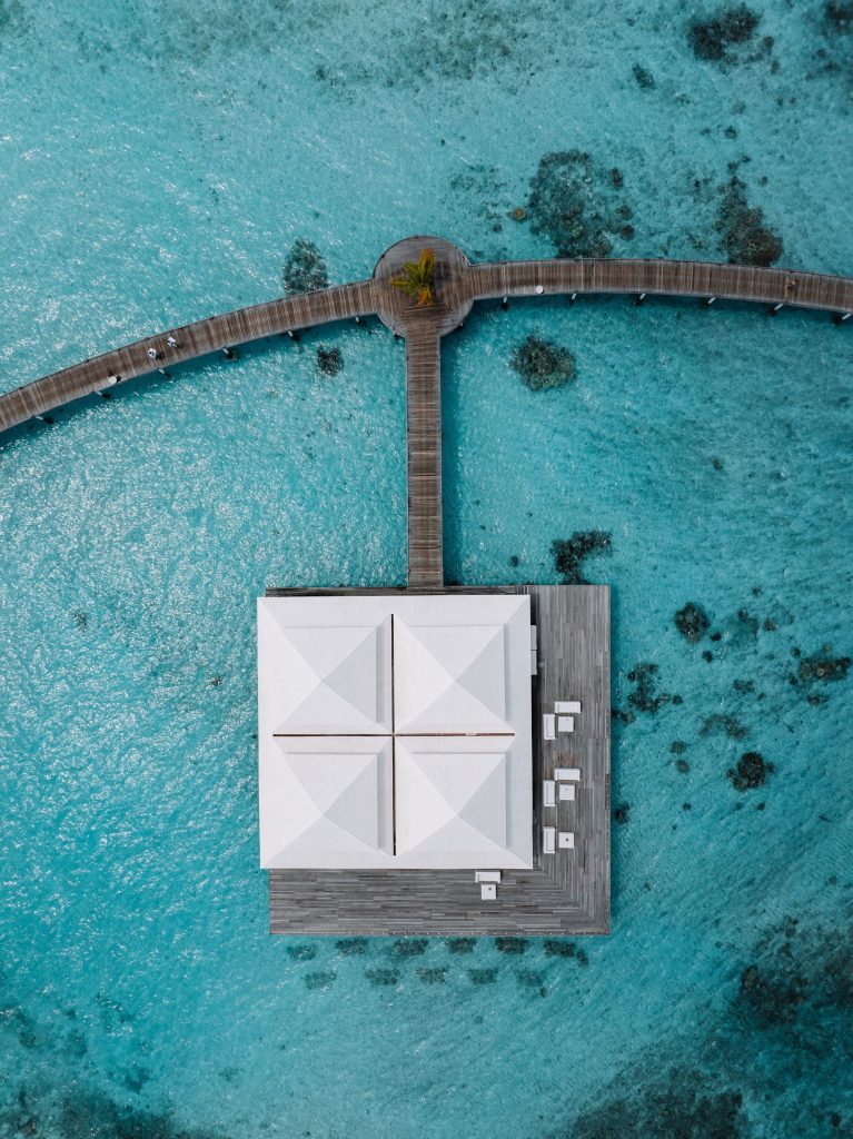 Aerial view of a white-roofed building on a wooden pier over clear blue water, connected by a curved walkway with small patches of coral visible beneath the surface.
