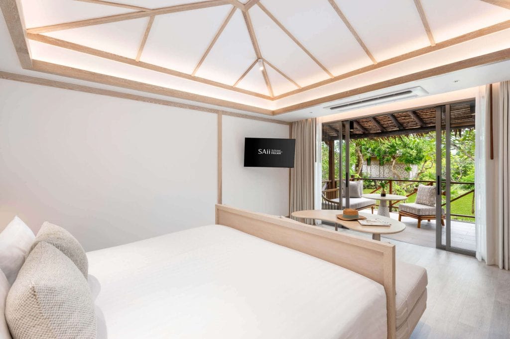 A bright, modern hotel room features a large bed, wall-mounted TV, and sliding glass doors opening to a private terrace with outdoor seating and lush garden views. The room has light wood accents and soft, neutral colors.