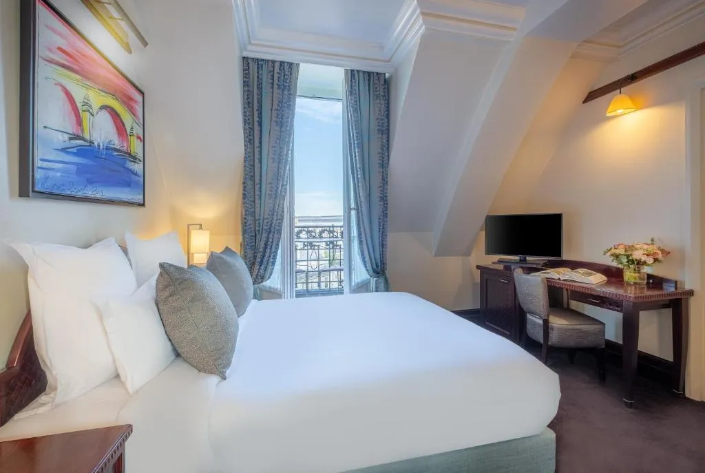 A bright hotel room with a large bed, two gray pillows, a colorful painting above the headboard, a desk with a TV and flowers, and tall windows with blue curtains overlooking a balcony and city view.