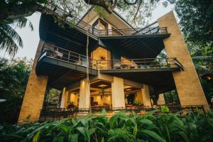 A modern, multi-story house with wide balconies and warm lighting, surrounded by lush green plants and tall trees, viewed from a low angle. People are relaxing on the balconies.