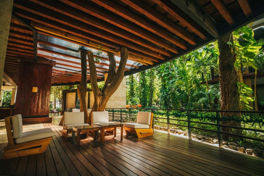 Outdoor patio with wooden floors and ceiling, modern white cushioned chairs, a small table, and lush green plants and trees surrounding the area, creating a relaxing and inviting atmosphere.