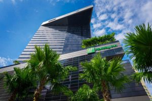A modern high-rise Holiday Inn hotel with a glass facade is seen from below, surrounded by tall palm trees under a blue sky with scattered clouds.