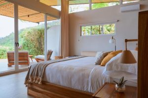 A cozy bedroom with a large bed, neutral bedding, and pillows. Floor-to-ceiling windows open to a patio with outdoor seating, offering views of greenery. Natural light fills the room, creating a warm, inviting atmosphere.