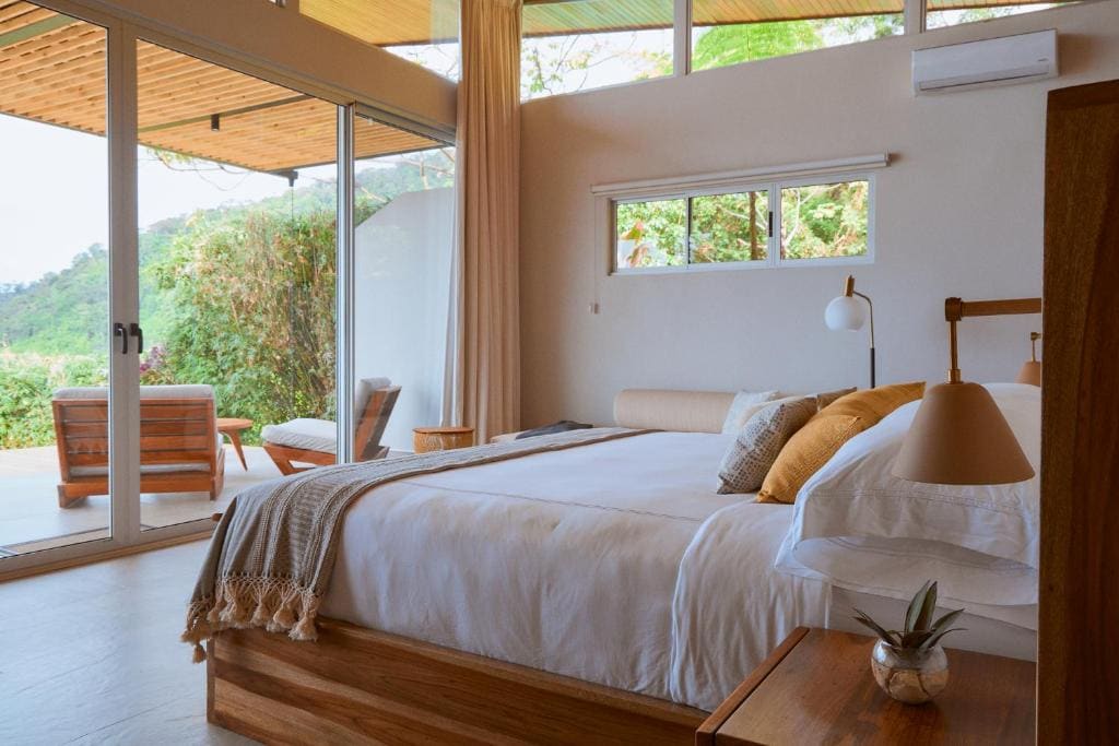 A cozy bedroom with a large bed, neutral bedding, and pillows. Floor-to-ceiling windows open to a patio with outdoor seating, offering views of greenery. Natural light fills the room, creating a warm, inviting atmosphere.