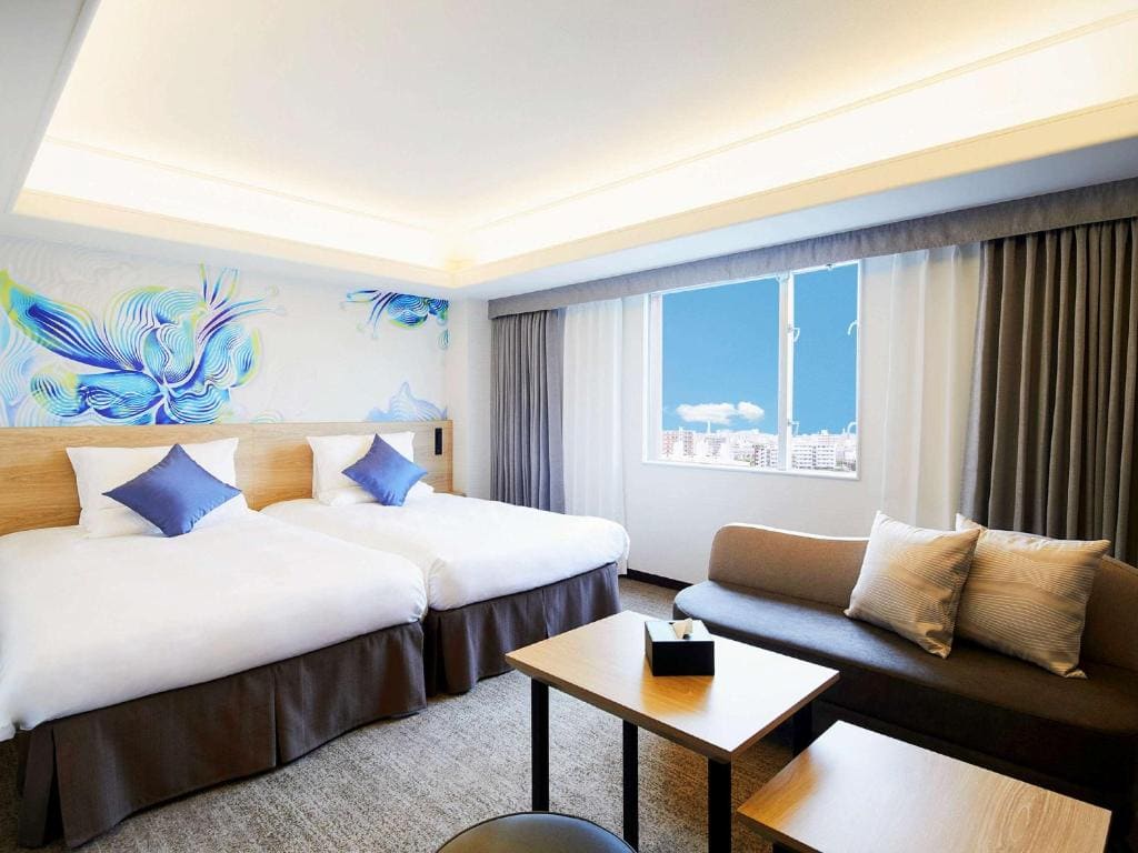 Modern hotel room with two twin beds, blue accent pillows, a wall mural with abstract floral design, a sofa with cushions, coffee table, and a large window with city view and blue sky.