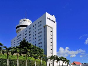 A tall white Novotel hotel building with a round observation deck rises above palm trees and greenery under a clear blue sky with a few scattered clouds.