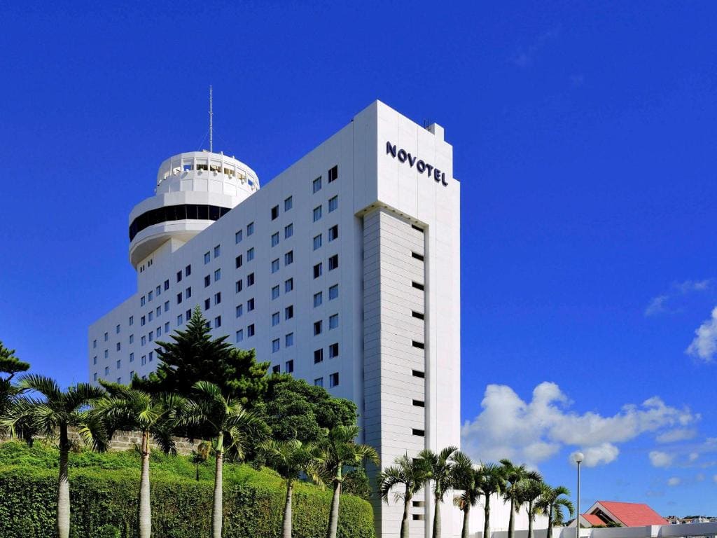 A tall white Novotel hotel building with a round observation deck rises above palm trees and greenery under a clear blue sky with a few scattered clouds.